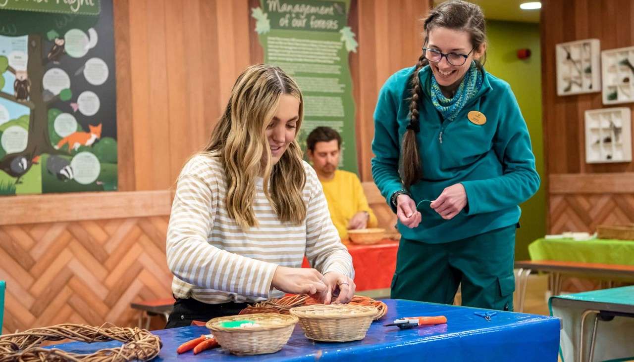 Woman weaves wicker strands at a table, guided by an instructor, in a craft workshop. Baskets, tools, and materials sit nearby; another person works behind them. Wall posters include the headline: Management of our forests.