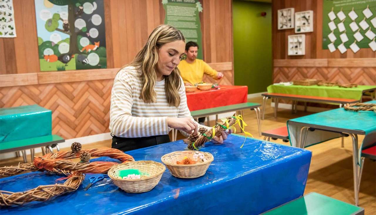 Woman weaves a twig wreath, tying ribbons and pinecones; in a craft workshop room with plastic-covered tables, baskets of supplies, another participant behind, and nature-themed posters on wooden walls.
