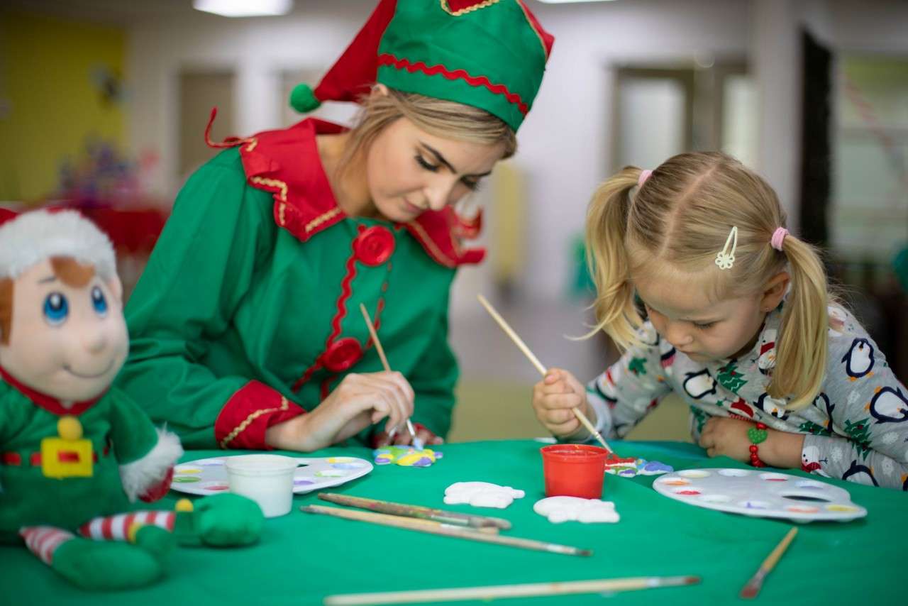 Four people dressed as Christmas elves dance with hands on heads in a festive room, wood-paneled backdrop with toys, wreaths, candles, and garlands.