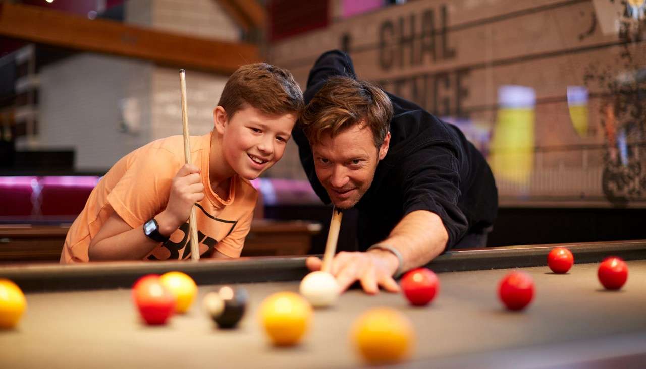 Man and boy play pool; man aims the cue at a white ball while boy watches closely. In a bar-like setting with red seats; background wall shows the words "CHAL" and "LENGE".