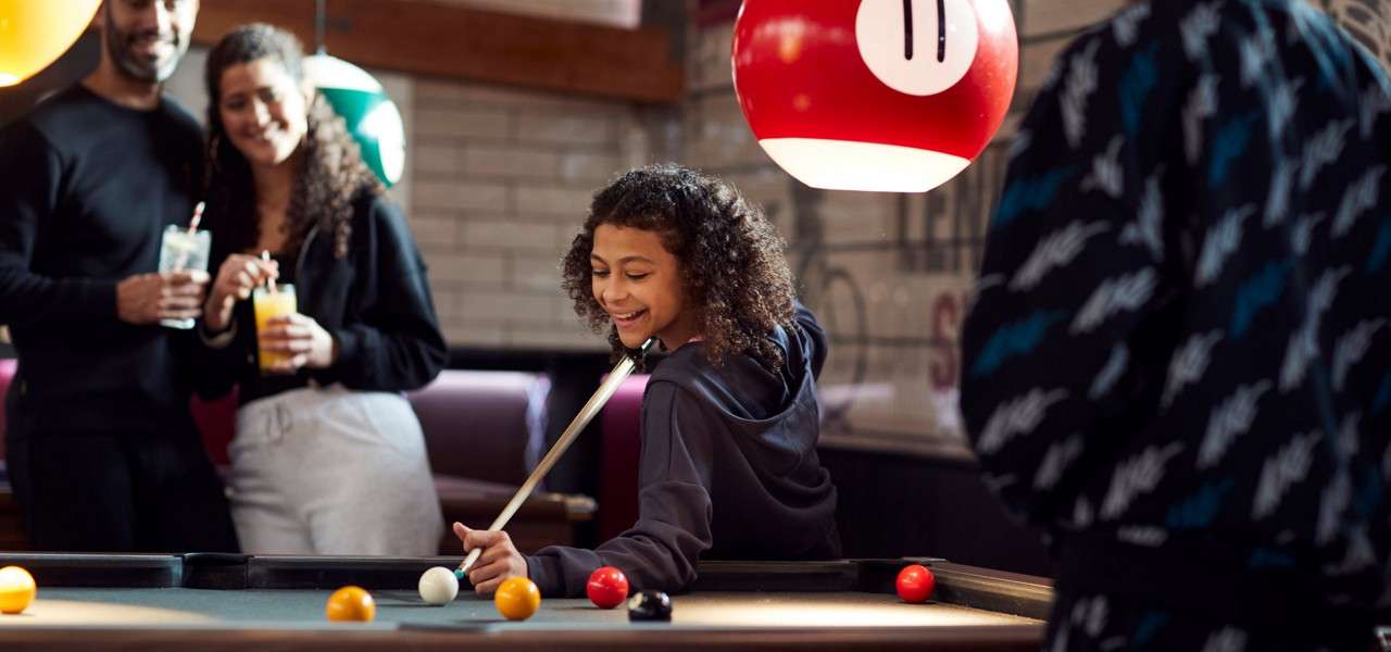 Young girl playing pool surrounded by her family.