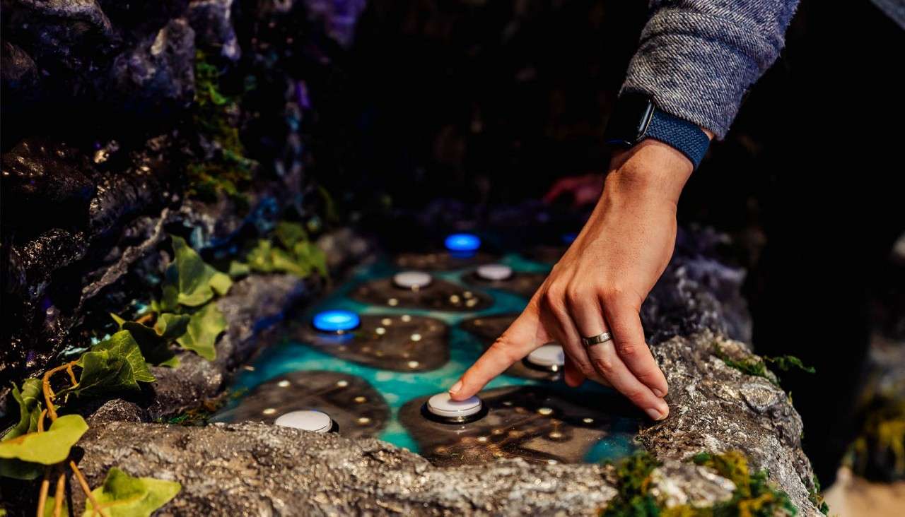 Hand presses a round button on a rocky, themed control panel with glowing blue and white lights, surrounded by artificial moss and vines, suggesting an interactive exhibit or game.