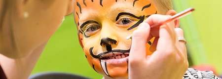 Child’s face—painted like a tiger—receives final brush strokes from an artist, close-up, in an indoor setting with a bright green background.