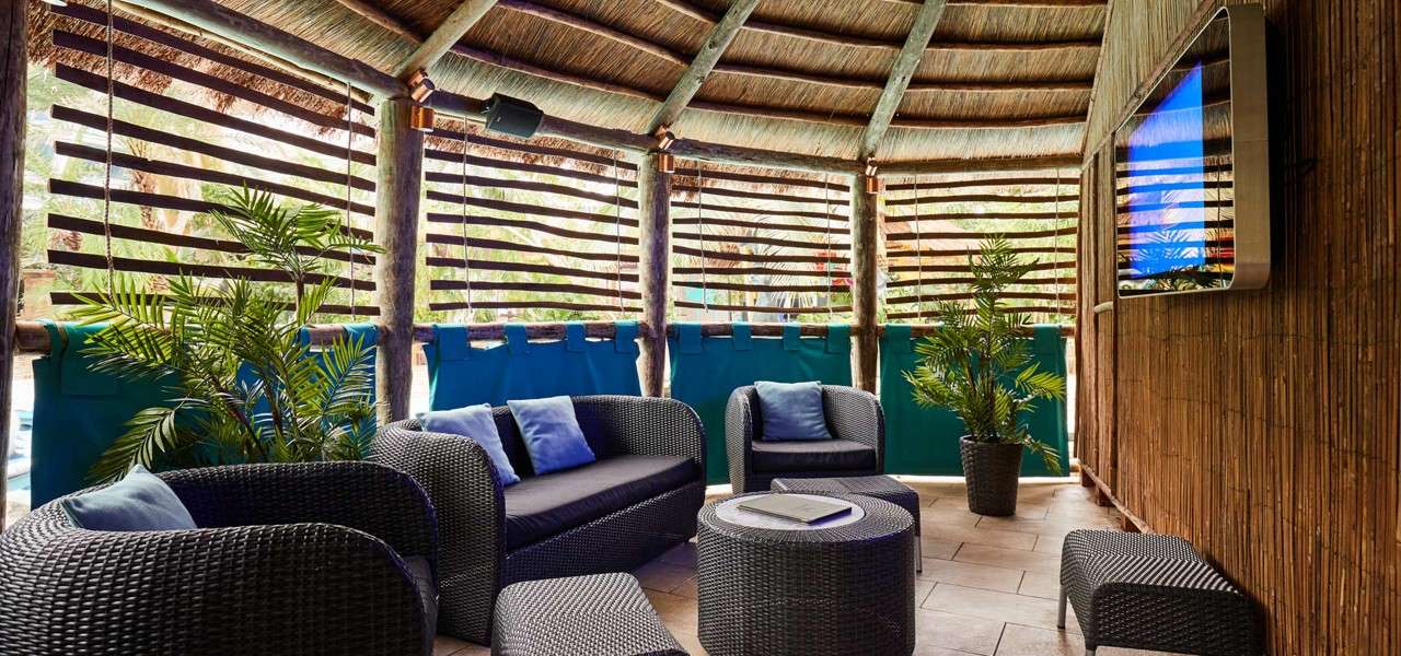 Wicker lounge furniture with cushions circles a small table, set under a thatched-roof cabana; potted palms, slatted walls, teal panels, and a wall-mounted TV create a shaded, tropical seating area.