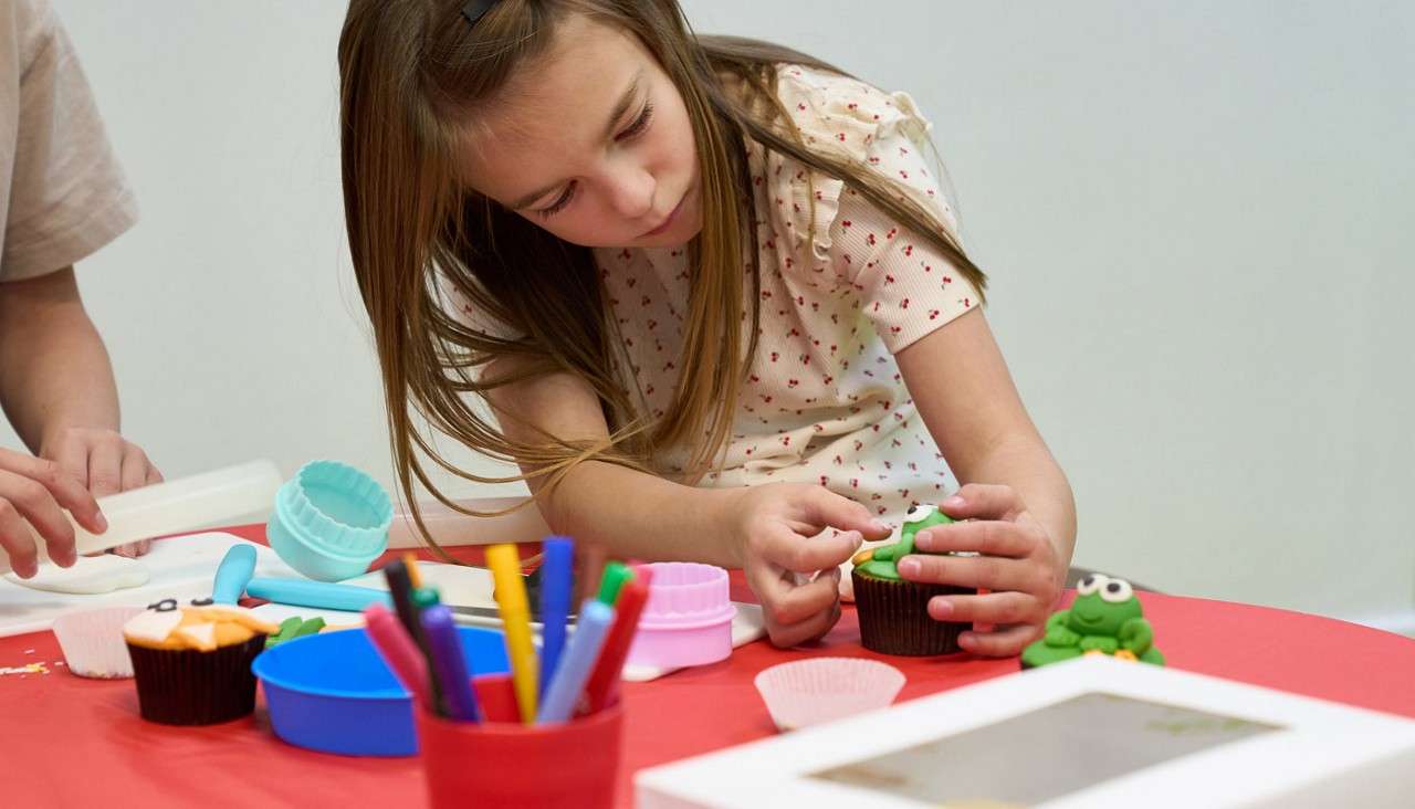 Child decorates a cupcake, pressing green fondant details with focused hands. On a red table, baking tools, silicone cutters, markers, and paper liners surround her; another person rolls fondant nearby.
