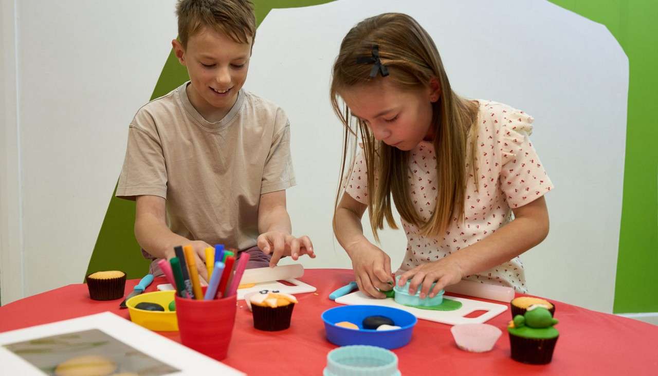 Two children press colored clay with cutters and shape decorations on small boards, surrounded by markers, bowls, and cupcakes, at a red table in a bright, white-and-green room.