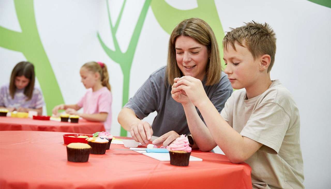 Children and an adult decorate cupcakes, placing fondant and icing on top. They sit at red-covered tables in a bright classroom with green wall graphics; bowls of toppings surround them.