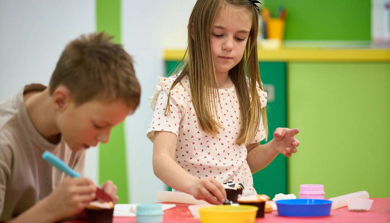Two children decorate cupcakes; one pipes frosting, the other places a wrapper on a cupcake. They work at a red table with colorful bowls and tools in a green-walled classroom.