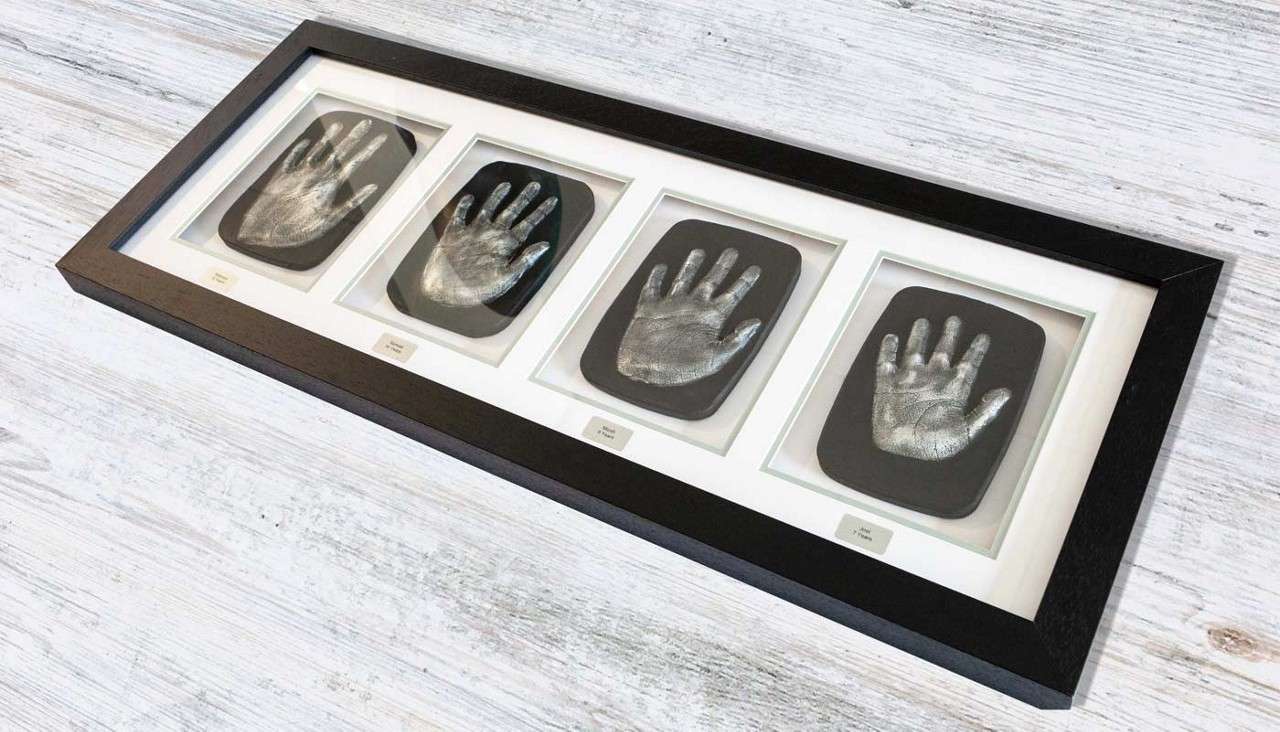 Four metallic hand impressions sit mounted in a long black frame, each on a dark plaque with small illegible labels, resting on a light wood surface, arranged side-by-side for display.