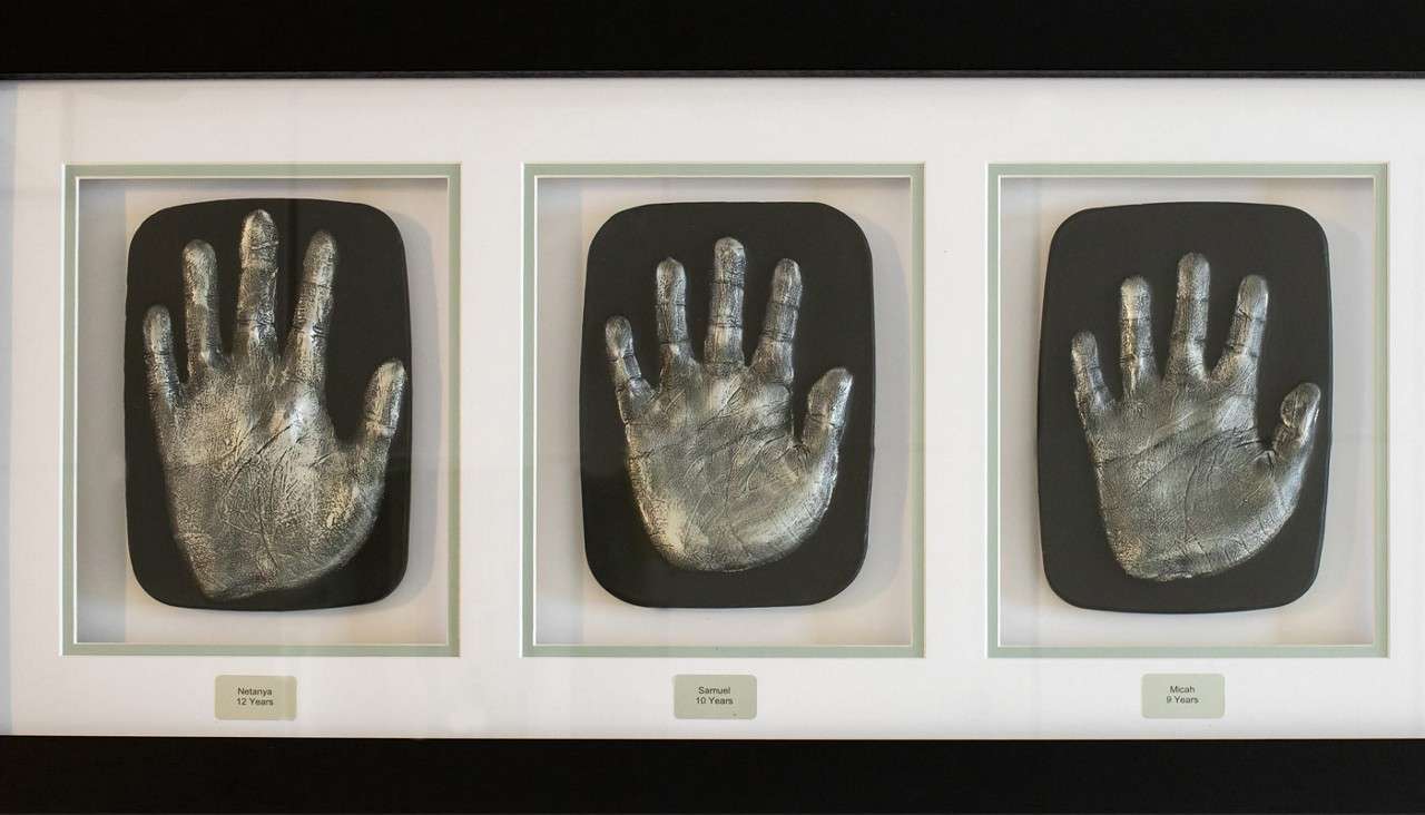 Silver handprint casts displayed side-by-side, mounted on black plaques in a white triple frame; labels read: Natanya 12 Years, Samuel 10 Years, Micah 9 Years.