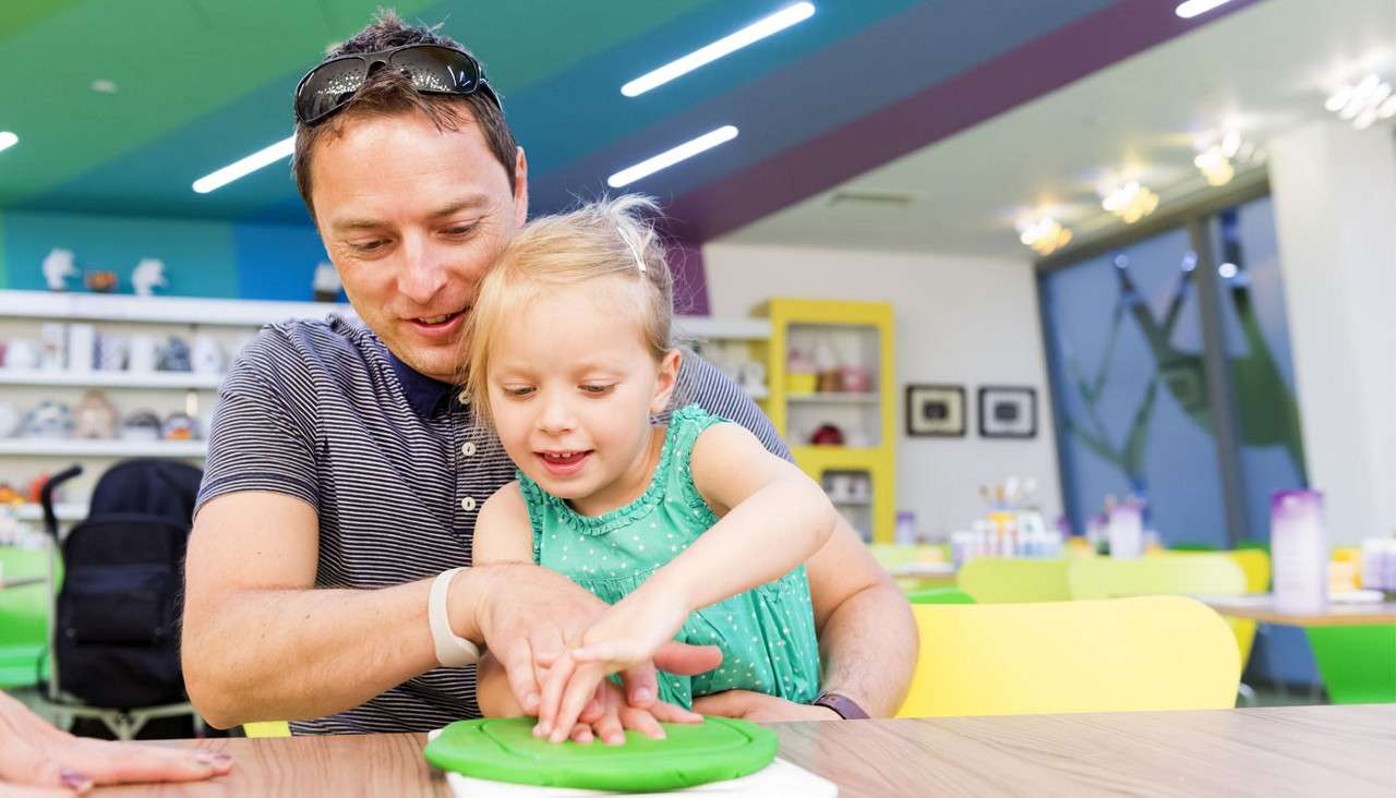Adult and child press hands into green clay, smiling gently, in a bright, colorful art studio with yellow chairs, shelves of craft supplies, and ceiling lights above large windows.