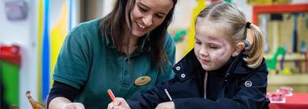 Adult and child draw with colored pens, the adult guiding the child, at a table in a brightly colored classroom or playroom.