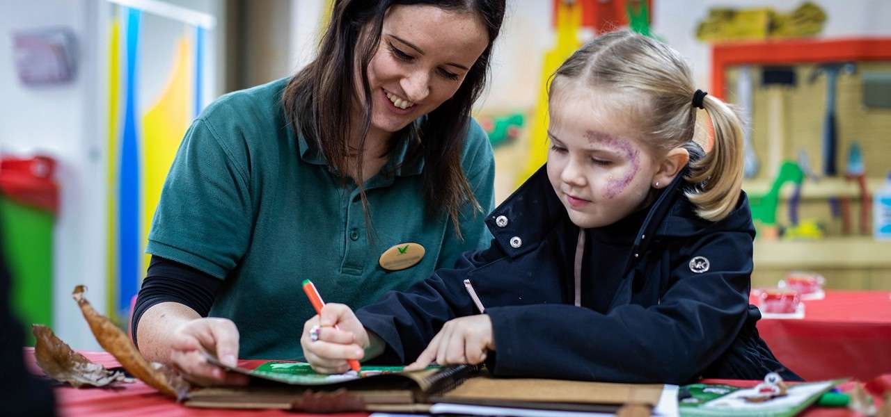 Child draws with a marker; an adult guides her hand. They sit at a red craft table with leaves, paper, and supplies inside a colorful classroom with shelves.