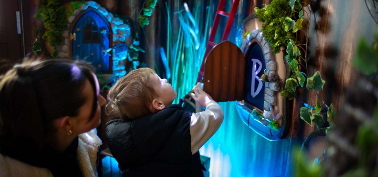 Child opens a small wooden door and peers inside, while an adult watches; set within a fantasy-themed, ivy-covered interactive wall glowing blue, with stone-framed windows and a sign reading "B".