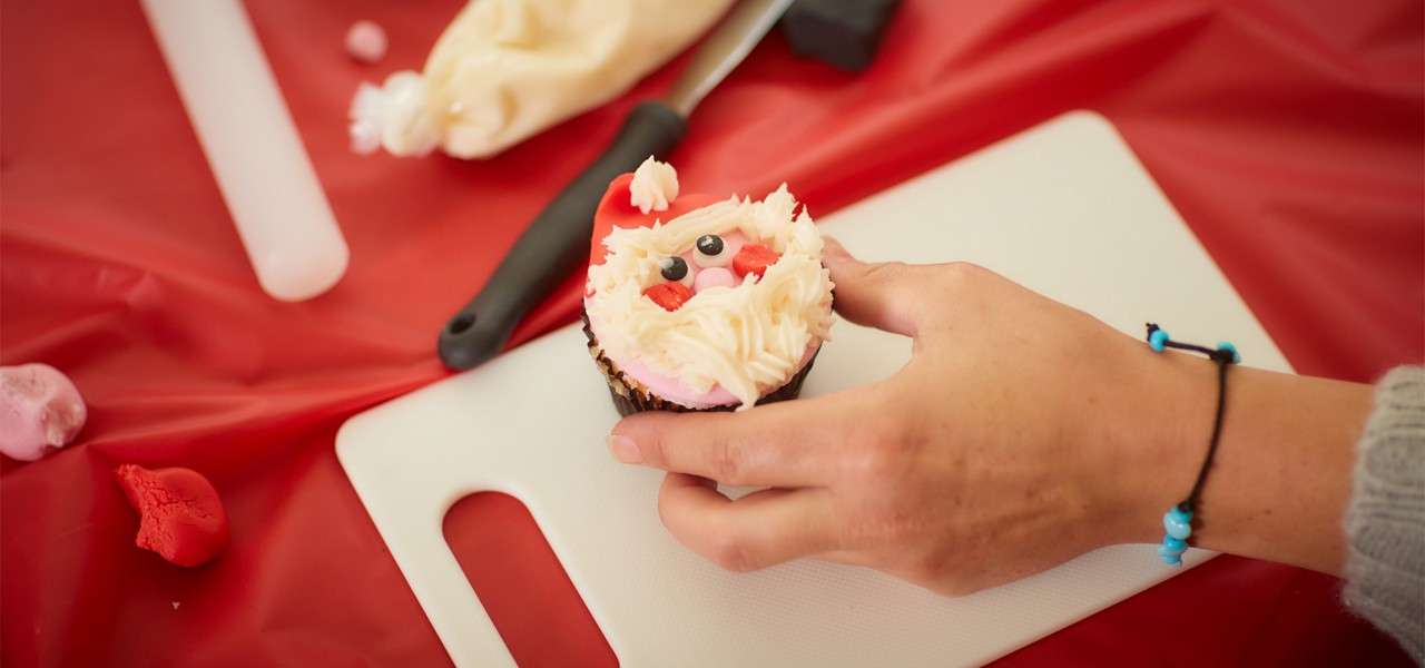 Santa-face cupcake is held by a hand, with piped beard and eyes. Nearby are a cutting board, piping bag, spatula, and red fondant scraps on a red table.