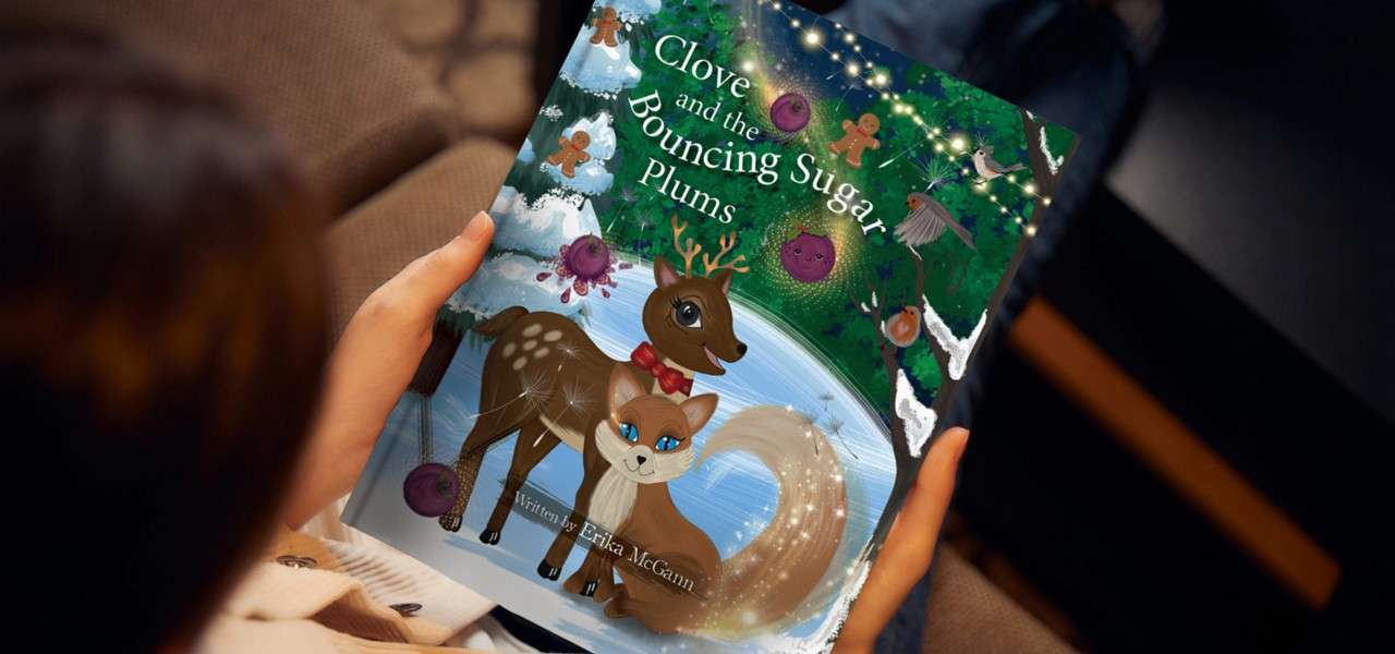 A children's book is held on a couch; its cover shows a deer and a fluffy-tailed animal near a snowy forest stream. Text: "Clove and the Bouncing Sugar Plums"; "Written by Erika McGann".