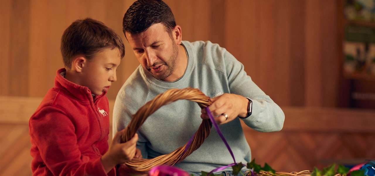 Adult and child weave a wicker wreath, threading a purple ribbon, at a table with greenery and ribbons in a warm, wooden indoor room.