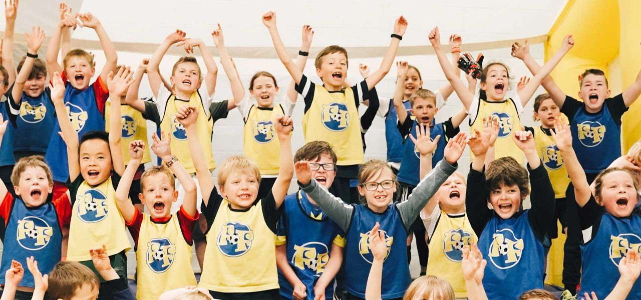 Children cheer with raised arms, wearing blue and yellow sports pinnies bearing a circular logo with stylized letters. They stand on indoor bleachers in a gym, smiling and celebrating together.