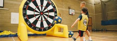 Giant inflatable dartboard awaits; a child and an adult approach with soccer-ball darts in an indoor gym. Visible text: numbers 17, 3, 19 on the board, and M on the child’s vest.