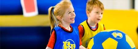 Smiling child lifts a large blue-and-yellow soccer ball, while another child watches; both wear blue training bibs over red shirts in a brightly colored indoor play area with inflatable barriers.