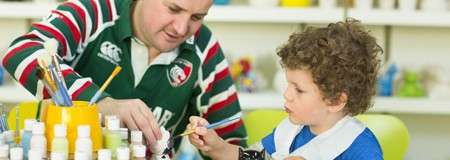 Child paints a small object while an adult assists, surrounded by paint bottles and brushes on a table in a bright classroom. Visible text: CCC, AR.
