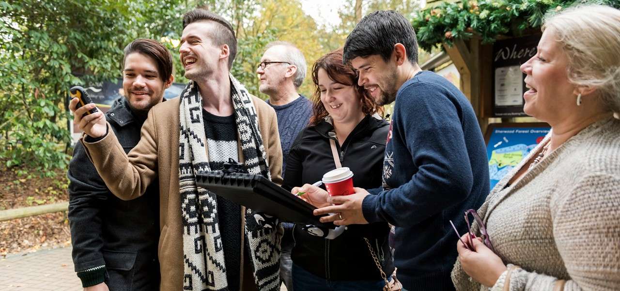 Group of adults laughing and collaborating, checking a rugged tablet and handheld device; outdoors at a park entrance with signboards, greenery, and one holding a red coffee cup. Visible text: “Where”.