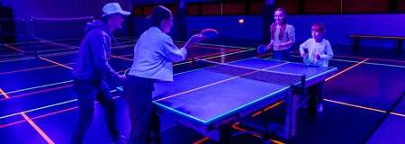 Four people play table tennis, two per side, swinging paddles. The game occurs in a dark indoor court lit by UV light and neon lines, with benches and badminton markings.