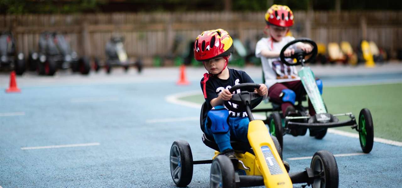 Two children steer pedal go-karts, wearing helmets and knee pads, racing around a marked outdoor track with cones, with a row of parked karts and a wooden fence in the background.