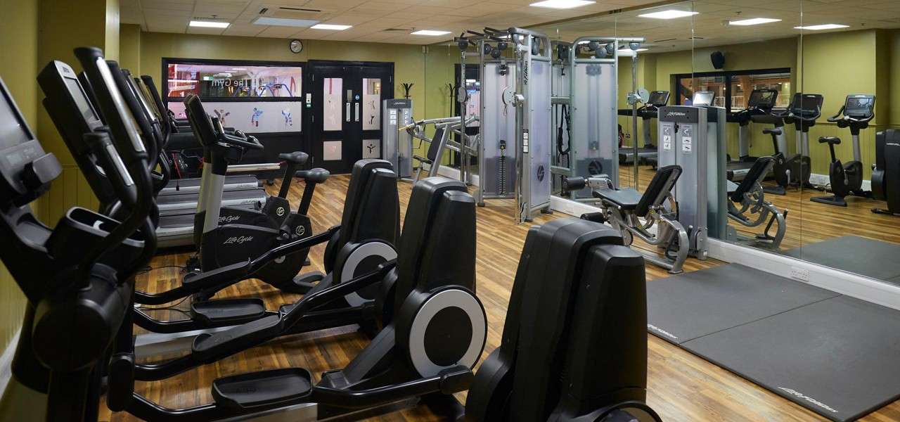 Cardio machines—ellipticals and bikes—stand idle, facing mirrors; weight machines line the side in a clean, wood-floored gym. Visible text: Life Fitness; The Gym.