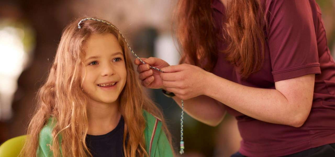Smiling child with long hair sits as an adult wraps a beaded strand into her hair; casual setting with soft, warm lighting and a blurred background.
