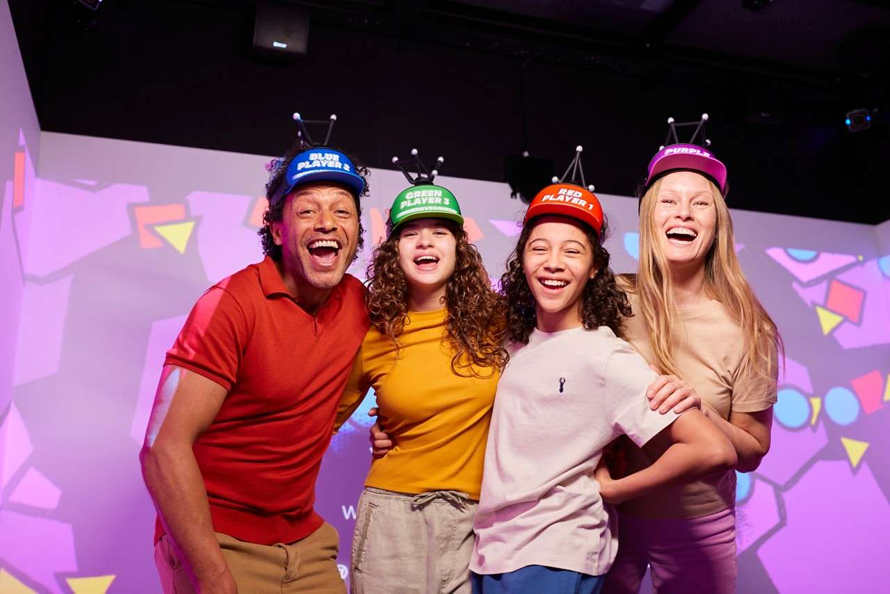 Four people wearing colored game caps pose smiling with arms around each other, in a room with projected geometric shapes. Visible cap text: BLUE PLAYER 2; GREEN PLAYER 3; RED PLAYER 1; PURPLE.