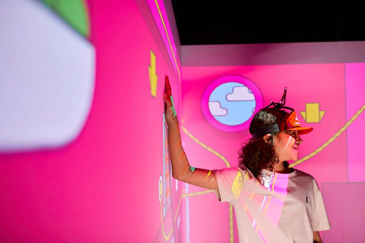 Person in a red cap with headgear touches a pink wall, interacting with projected cartoon graphics; round window shows clouds, yellow arrows and neon lines. Cap logo text is illegible.