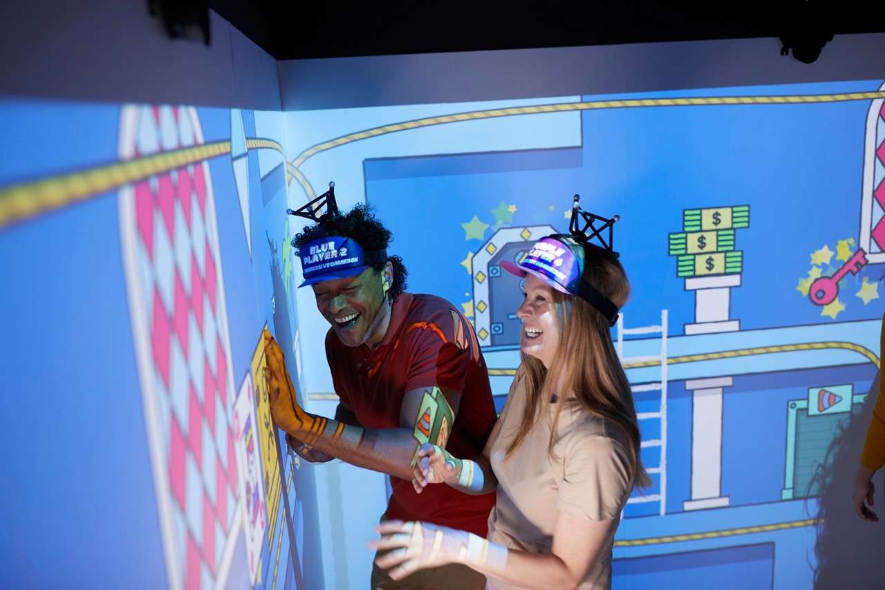 Two players wearing visor headsets touch and interact with a projected cartoon game wall in an immersive room. Text: “BLUE PLAYER 2”.