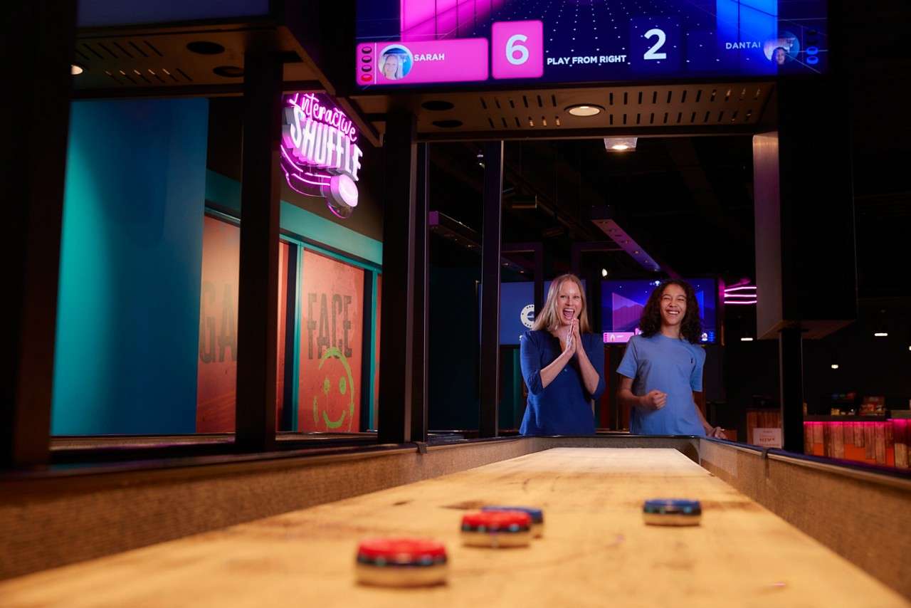 Shuffleboard pucks slide down a wooden lane as two people cheer in a neon-lit arcade; overhead scoreboard and sign glow. Text: interactive SHUFFLE; SARAH 6 PLAY FROM RIGHT 2 DANTAI; FACE.