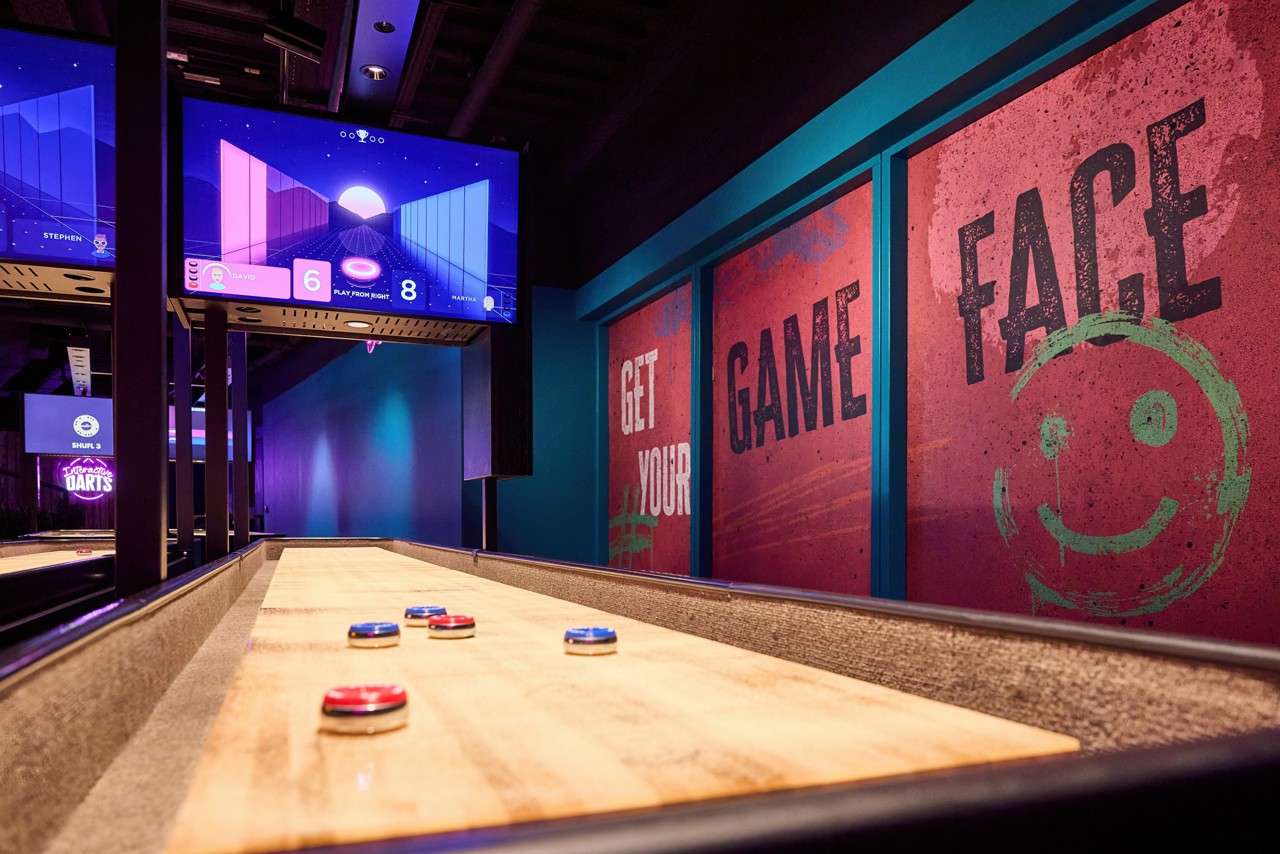 Shuffleboard pucks sit on a wooden lane, while overhead screens track scores in a neon-lit arcade bar. Text: GET YOUR GAME FACE; INTERACTIVE DARTS; SHUFL 3; PLAY FROM RIGHT; 6; 8; STEPHEN; DAVIS; MATTIA.