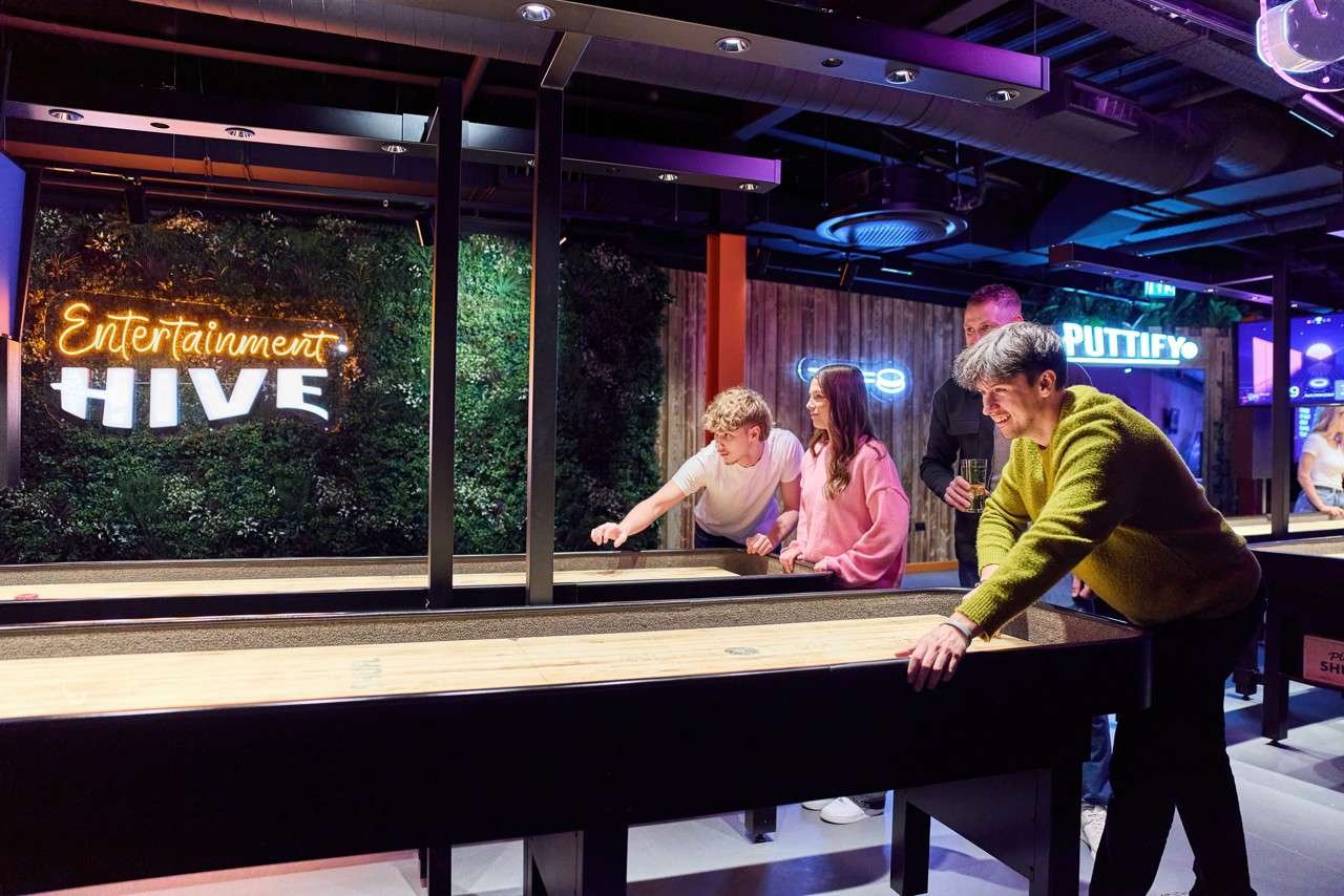 Players slide pucks down a shuffleboard table, smiling, in a neon-lit entertainment venue; wall signs read: “Entertainment HIVE” and “PUTTIFY.”