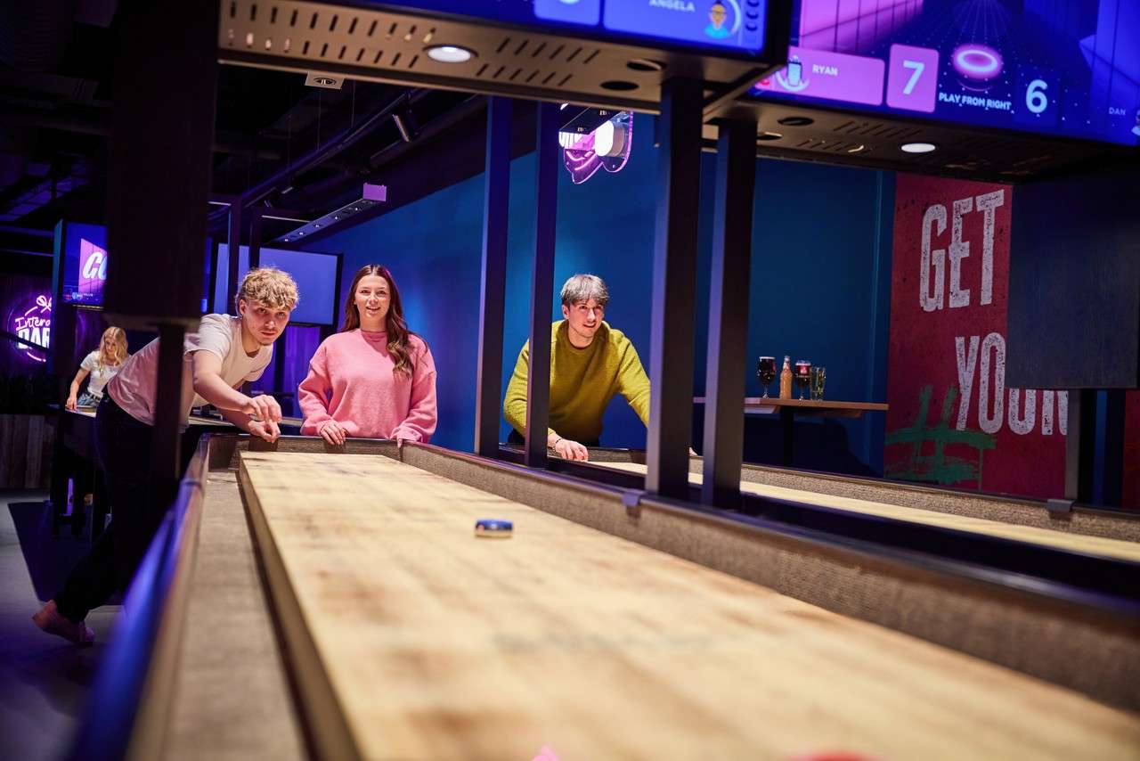 Shuffleboard players slide pucks along a long wooden lane in a neon-lit arcade bar. Text: RYAN 7; DAN 6; PLAY FROM RIGHT; ANGELA; GO; GET YOU.