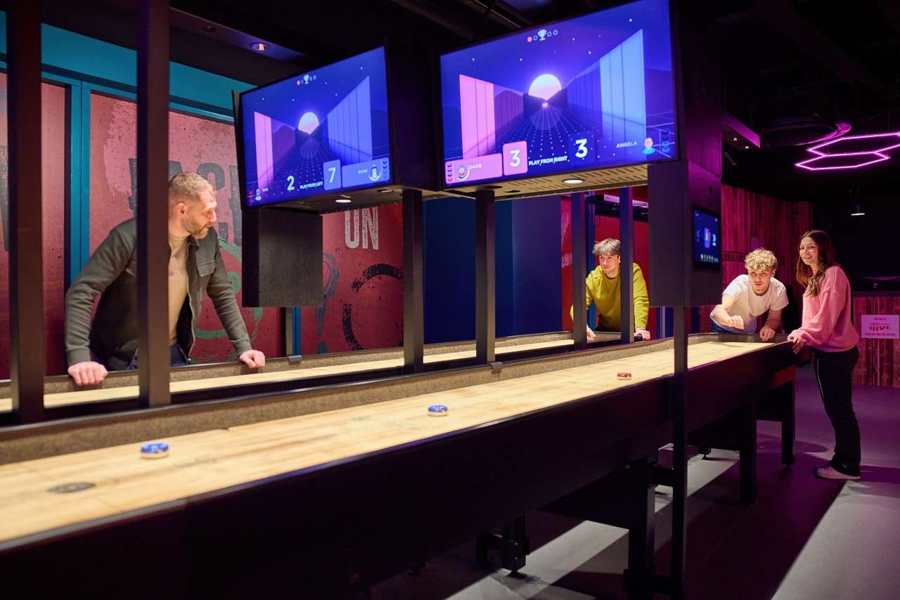 Players slide pucks on a shuffleboard table, competing beneath two score screens in a neon-lit arcade bar. Text: 2, 7, 3, 3, ON.