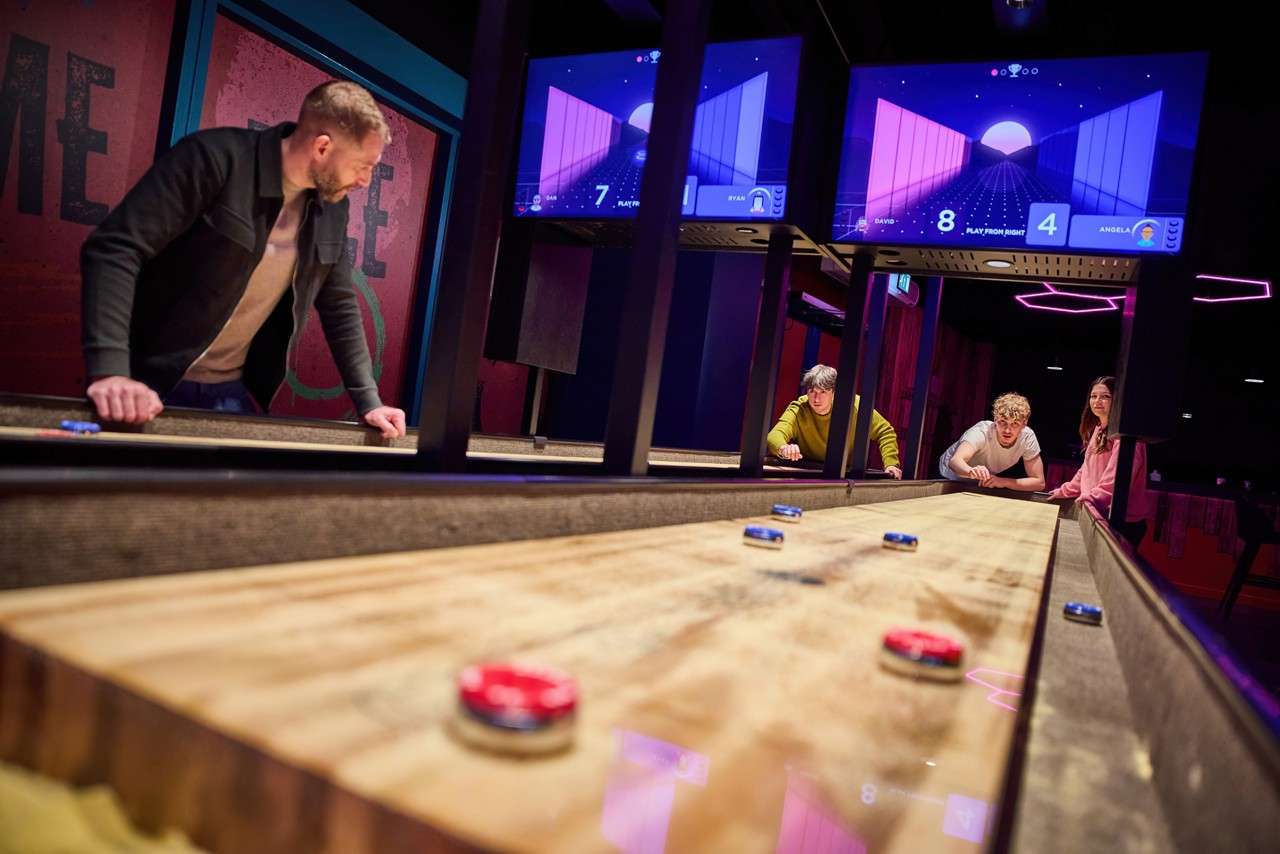 Shuffleboard pucks slide along a long wooden table as four people play, in a neon-lit arcade; overhead screens display: "7", "DAVID 8", "ANGELA 4", "PLAY FROM RIGHT", "RYAN", and wall text "ME".