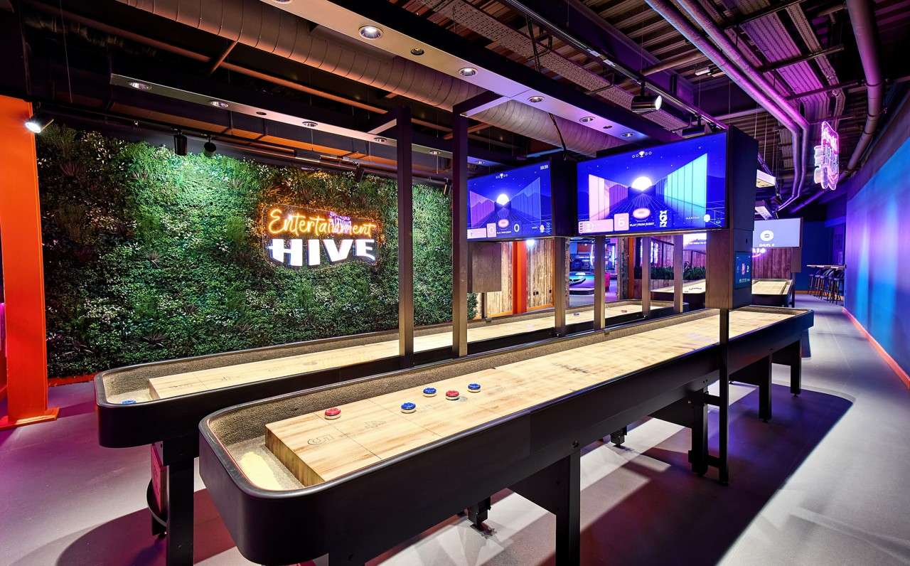 Shuffleboard tables await play under monitors; a plant wall bears neon words “Entertainment HIVE”; screens show the number “6” and other small, unreadable text.