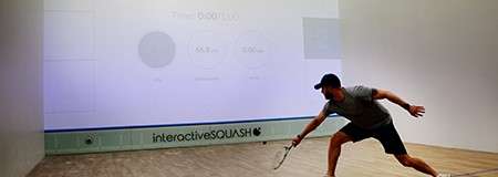 Squash player lunges low, swinging a racket toward the ball, on an indoor court where the front wall displays an interactive projection; branding along the base reads: interactiveSQUASH.