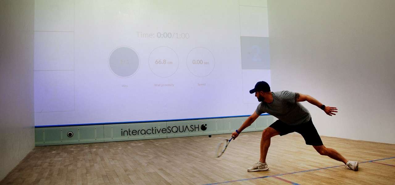Squash player lunges with racket, reaching for a shot; inside an interactive squash court with a projected display. Text: Time: 0:00/1:00; interactiveSQUASH.