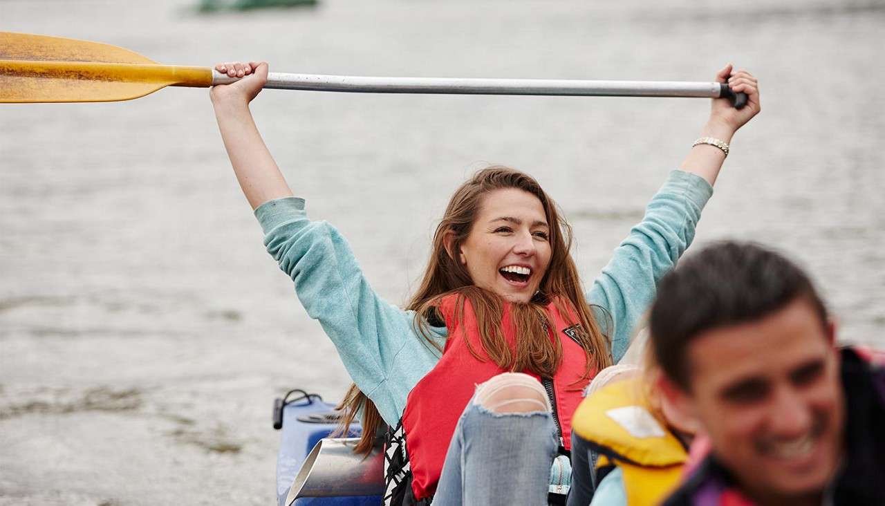 Kayaker lifts paddle overhead, laughing; she wears a red life vest and ripped jeans, seated in a blue kayak on calm water, with another paddler blurred in the foreground.