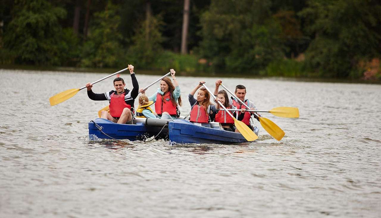 Six people in life vests paddle blue canoes together, raising oars mid-stroke, on a calm lake with a forested shoreline in the background.