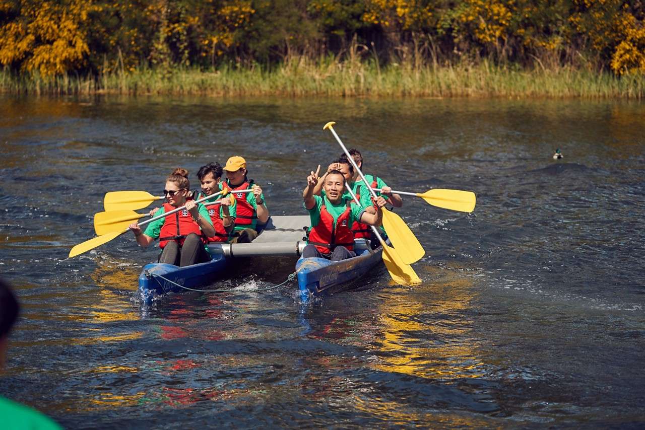 Group of people wearing red life vests paddle a twin-hulled blue canoe; a child sits between them; on a calm lake with a tree-lined shore.