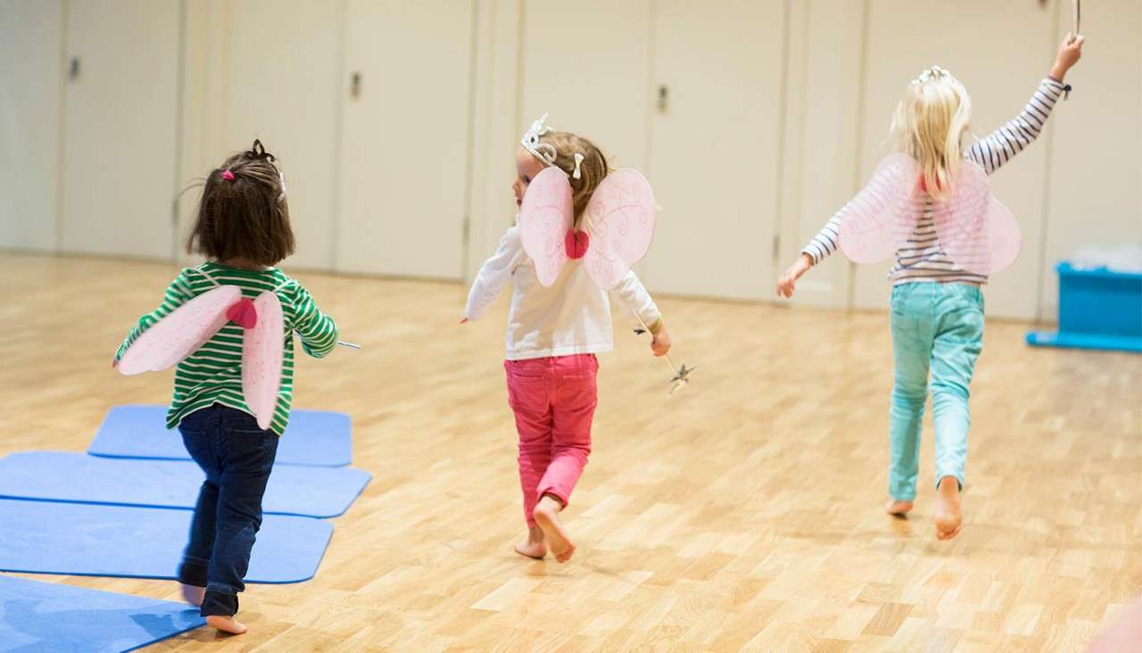 Three young children wearing pink fairy wings run barefoot, one holding a wand, across a wooden floor. They play in a bright indoor studio with blue mats and closed doors.