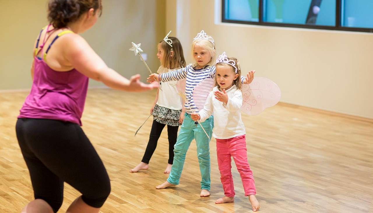 Three young children wearing tiaras and fairy wings hold wands and follow a teacher’s gestures, practicing movement on a wooden studio floor with large windows in the background.