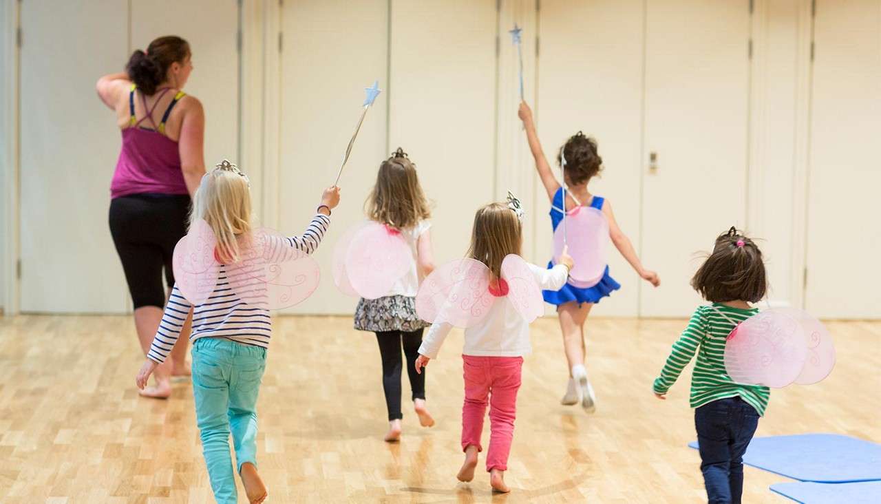 Children in pink fairy wings raise star wands and run barefoot after an adult instructor across a bright dance studio with a wooden floor and closed doors.