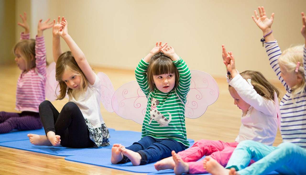 Children sit cross-legged on yoga mats, raising arms overhead, stretching together. In a bright studio with wood floor, one child wears fairy wings and a green striped shirt.