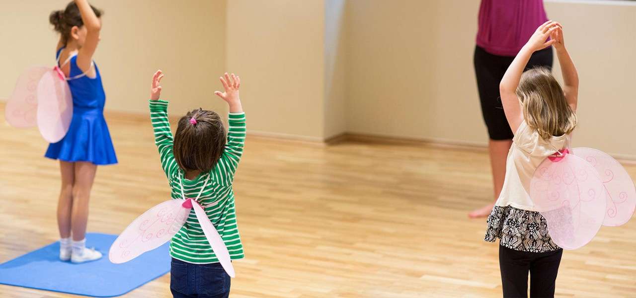 Three young children with pink fairy wings raise their arms while practicing dance poses, guided by an instructor, inside a bright wooden-floor studio; one child stands on a blue mat.